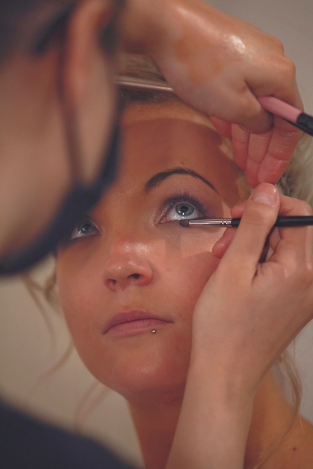 Makeup artist carefully applying eye makeup to blonde bride with two brushes.
