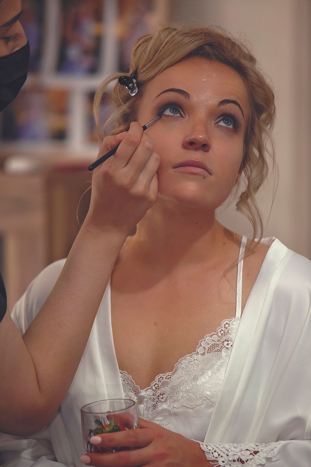 Bride in white lace robe holding drink while getting eye makeup applied.