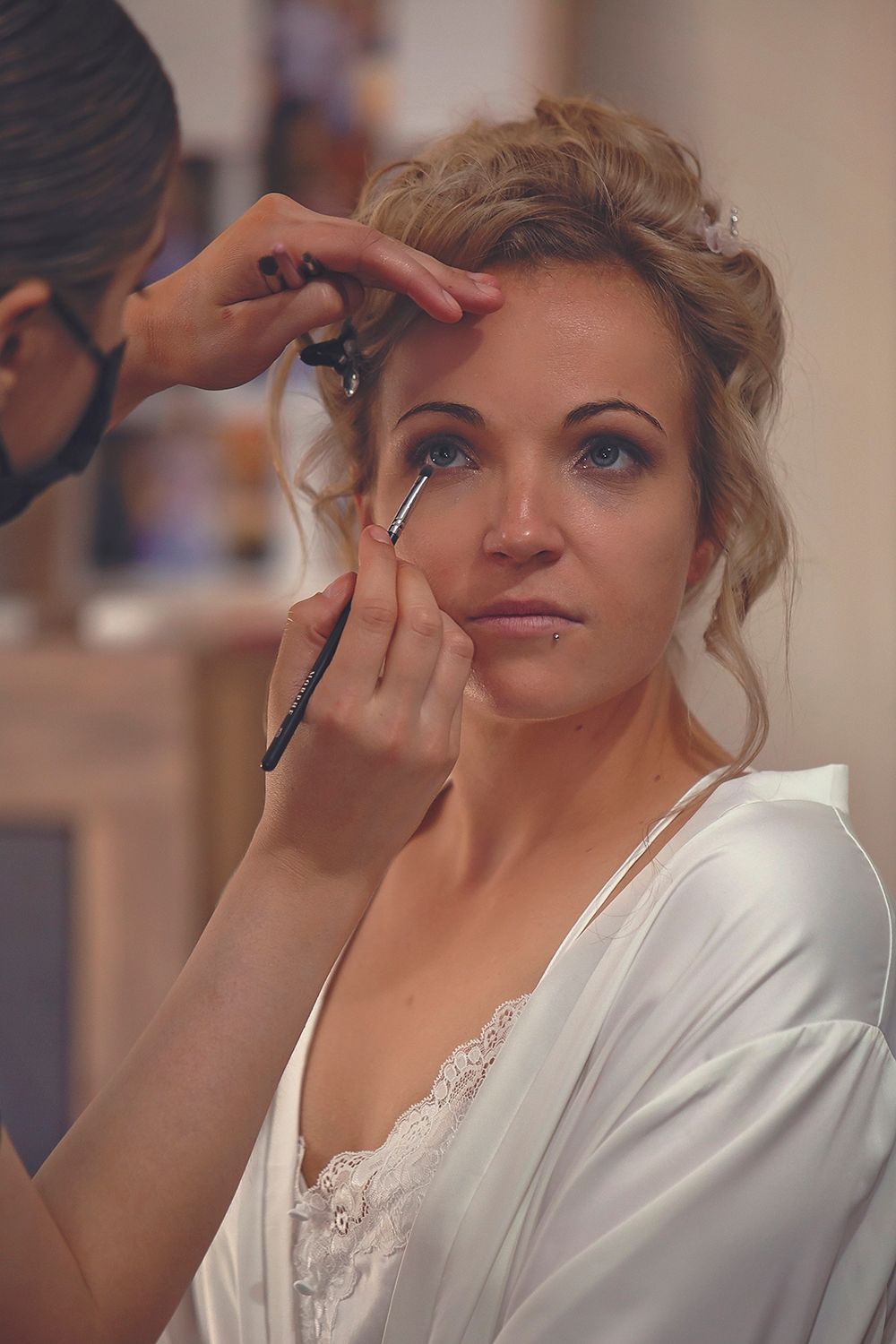 Makeup artist applying eye makeup to blonde bride in white lace robe.