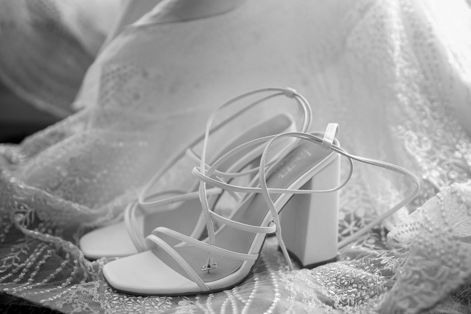 White strappy block heel wedding shoes on lace bridal gown in black and white photo.