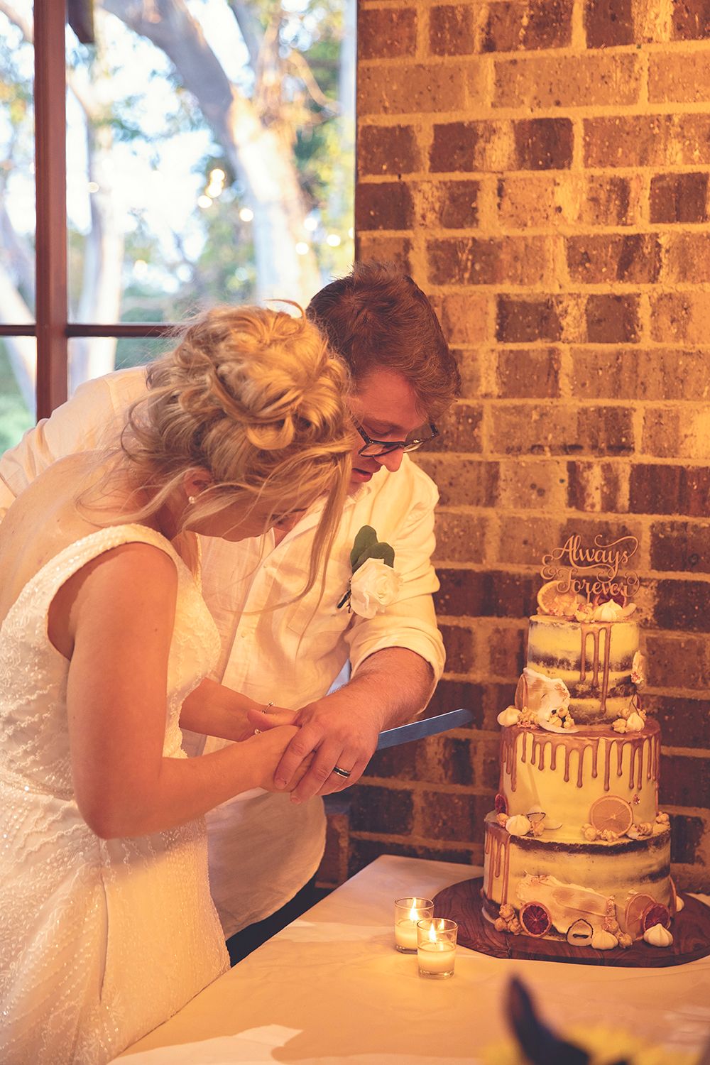Bride and groom cutting multi-tier naked wedding cake with candles against brick wall.