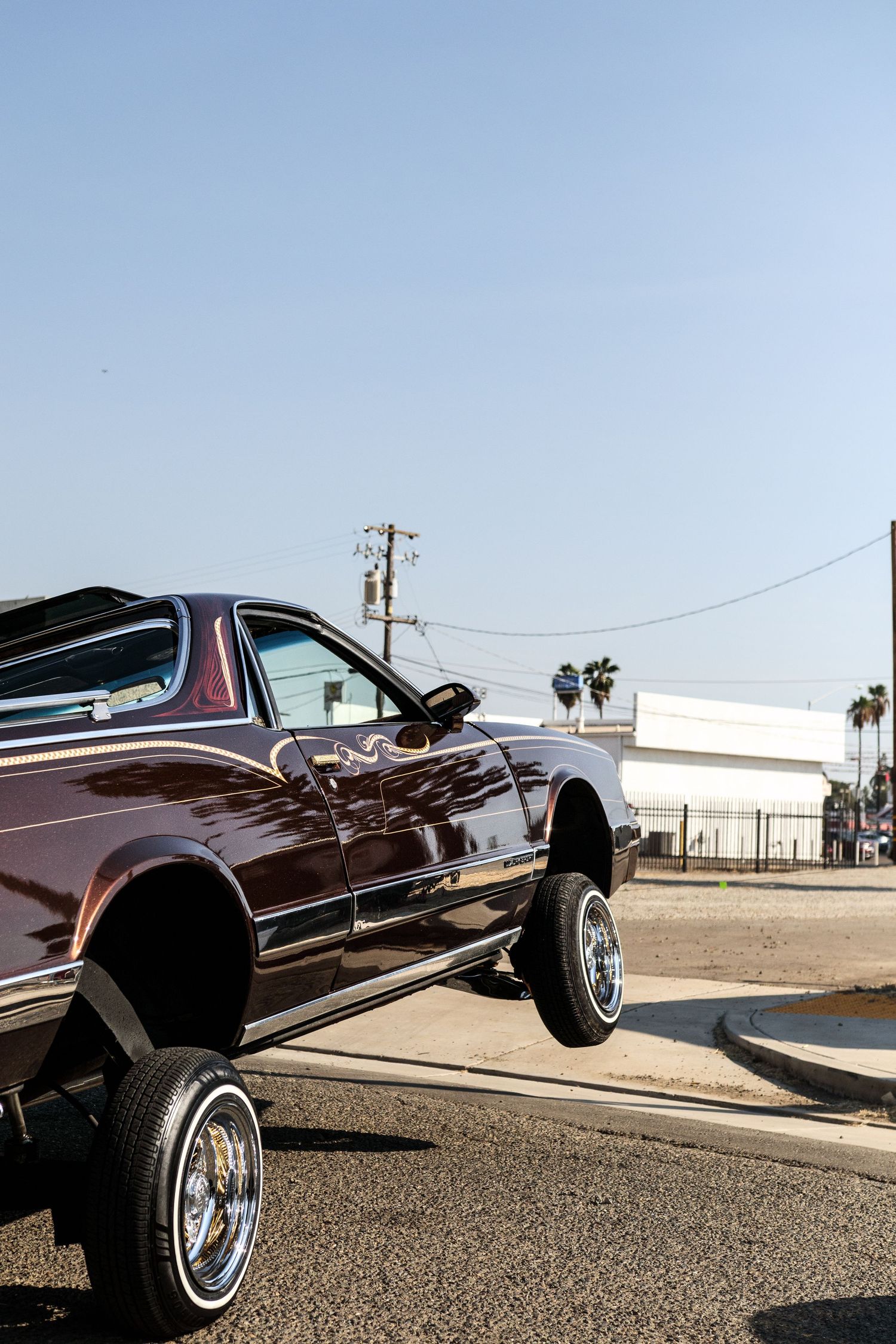 Shiny maroon lowrider car parked on a sunny street corner with palm trees nearby.