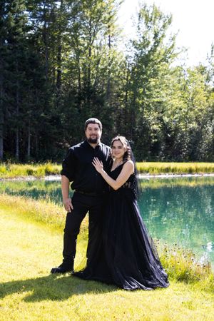 A couple in black formal attire poses romantically by a serene pond surrounded by lush green trees and grass.