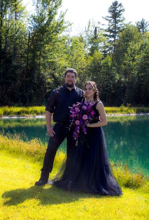 Couple in formal black attire poses by a vibrant turquoise lake surrounded by lush greenery and yellow flowers.
