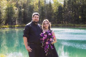 A couple poses by a turquoise lake surrounded by evergreen forest during a wedding photoshoot.