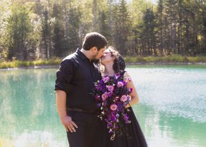 A couple shares a romantic moment beside a turquoise lake while holding a purple floral bouquet.
