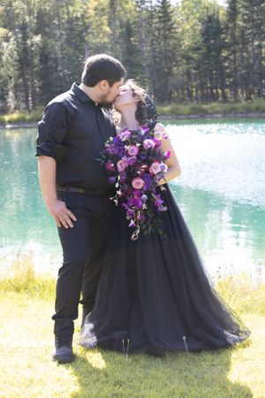 Wedding couple in black attire with purple flowers pose near a scenic mountain lake.