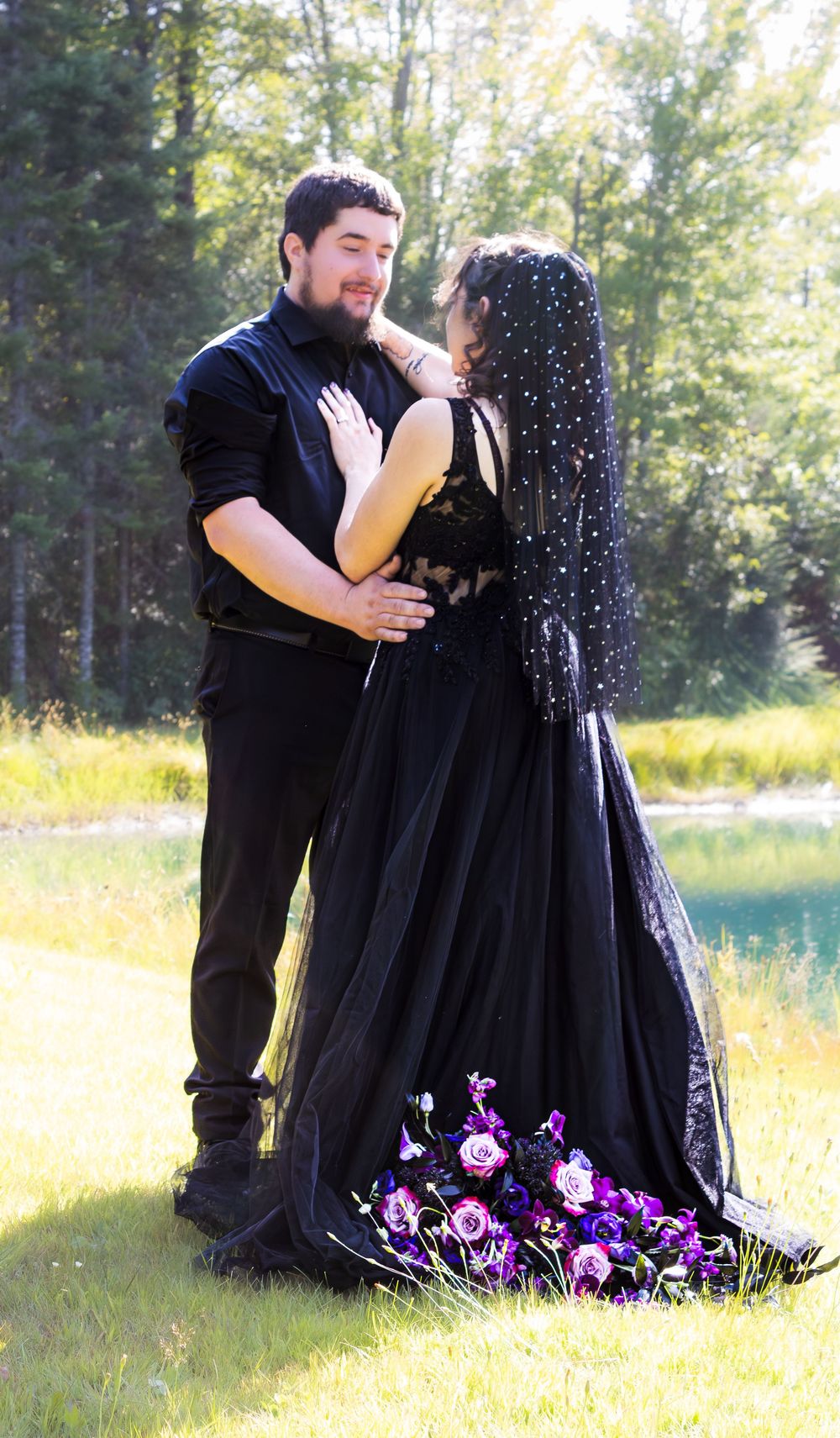 A couple in black formal attire embrace romantically in a sunlit meadow.