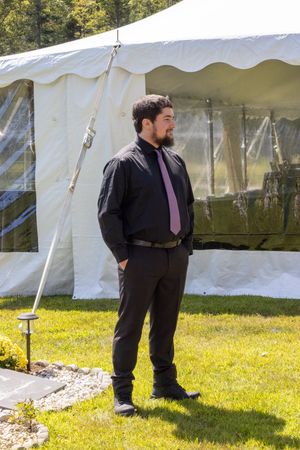 Person in formal black attire stands near white event tent on sunny grass lawn.