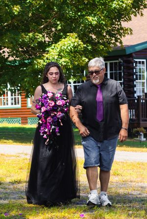 Wedding party members in black and purple attire walk across sunny outdoor venue.