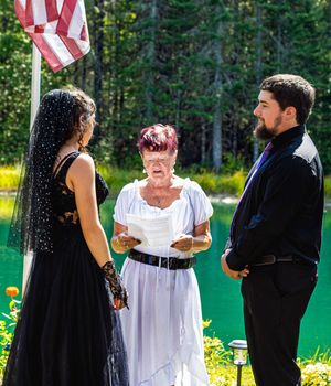 An outdoor wedding ceremony takes place near a lake with an American flag displayed.
