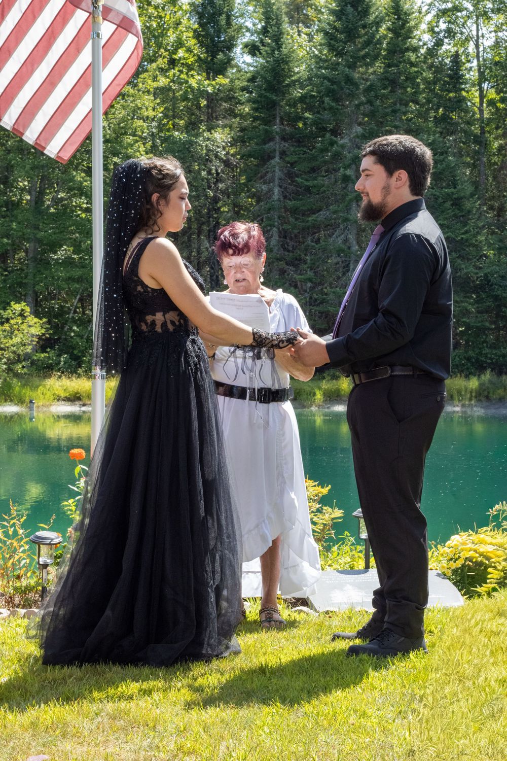 A wedding ceremony takes place by a turquoise lake with an American flag and evergreen trees in the background.