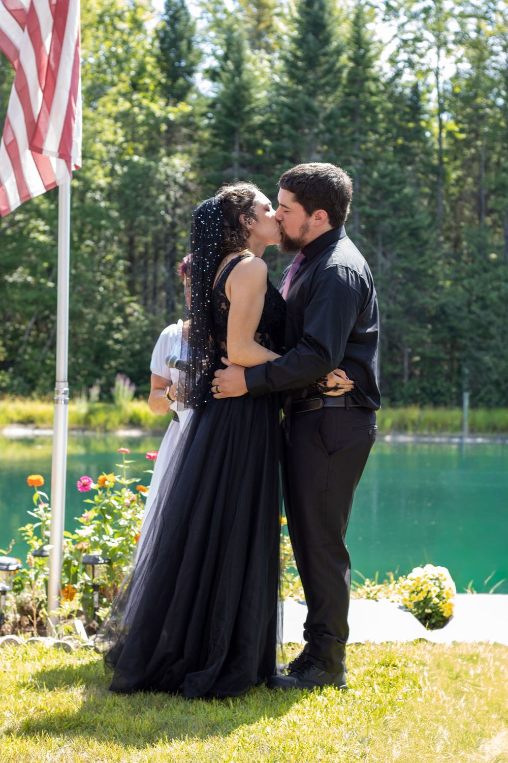 A couple shares a romantic kiss by a serene lake with evergreen trees and an American flag in the background.