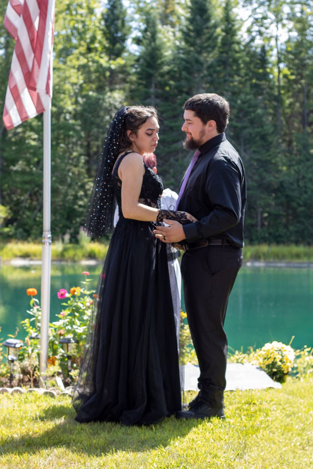 A couple in formal attire shares a tender moment on a grassy lawn with American flags and a lake in the background.