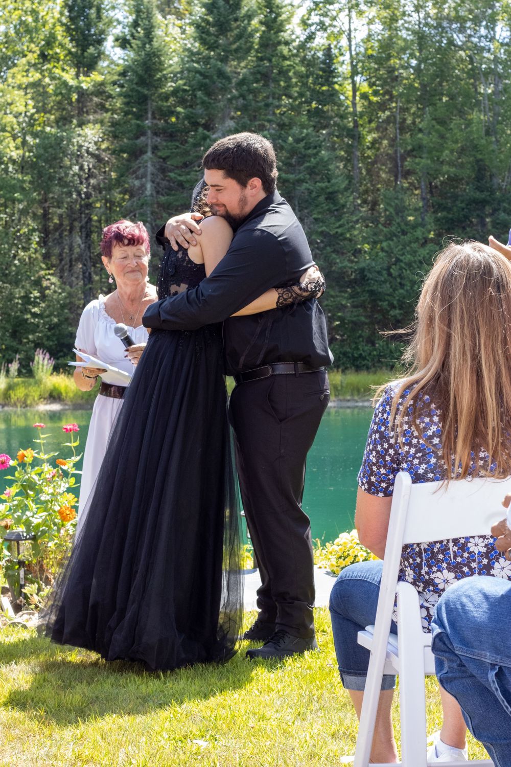 A couple embraces during an outdoor ceremony in a garden setting with white chairs and evergreen trees.