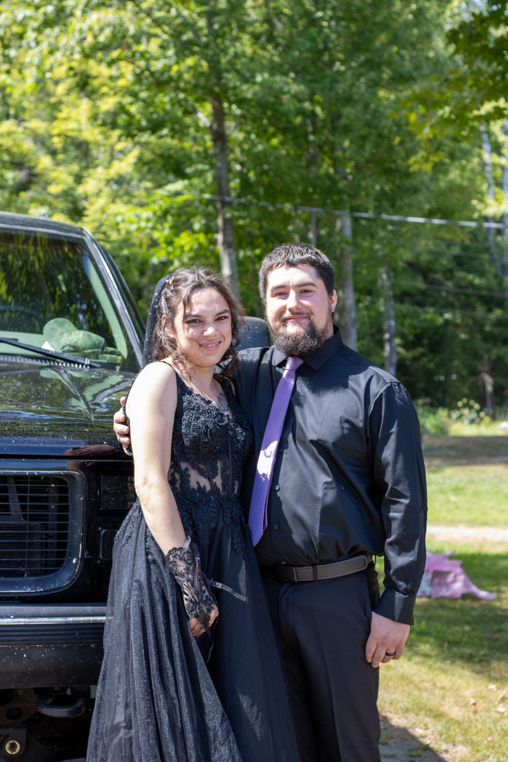 A couple in black formal wear poses together outdoors on a sunny day with trees and a car in the background.