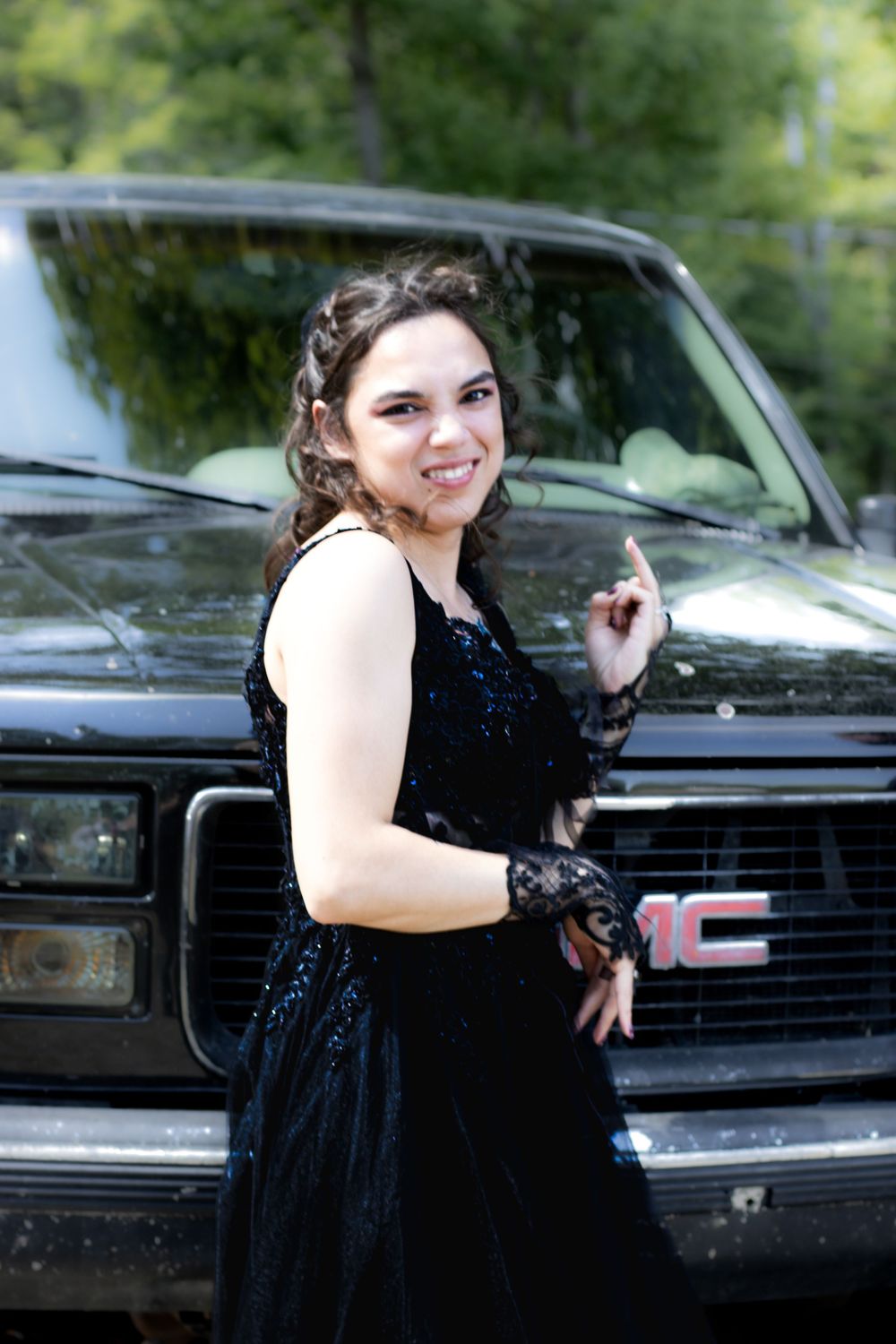 A person in a black formal dress poses playfully in front of a dark GMC truck outdoors.