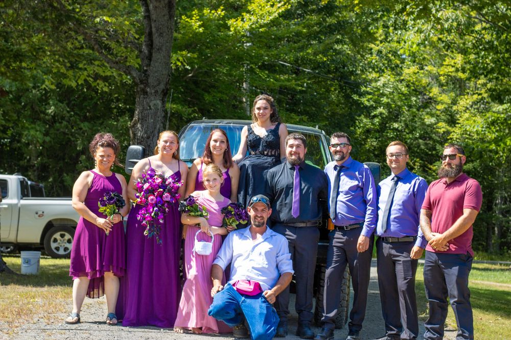Wedding party members in purple and blue attire pose together outdoors in a park setting.