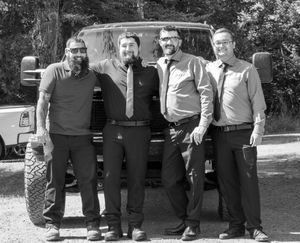 A black and white vintage photograph shows a group of people standing together in front of an old car outdoors.
