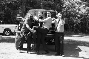 A group of people gather around a vehicle in a wooded outdoor setting during what appears to be a roadside stop.