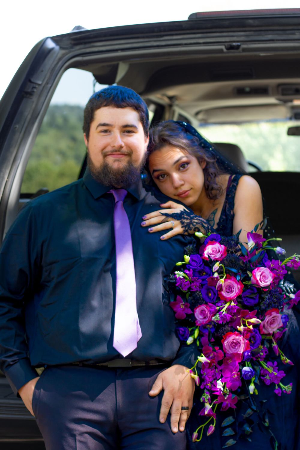 A couple poses inside a car with a vibrant purple and pink floral bouquet.