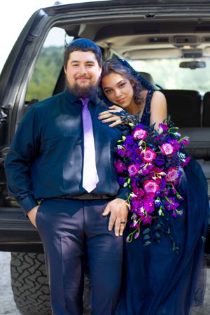 A couple in formal navy attire poses with vibrant purple floral bouquet inside a vehicle.