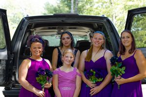 Wedding party members in purple dresses pose with bouquets in front of a black SUV.