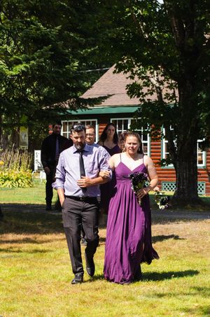 Wedding attendants walk across a sunny lawn in front of a rustic cabin.