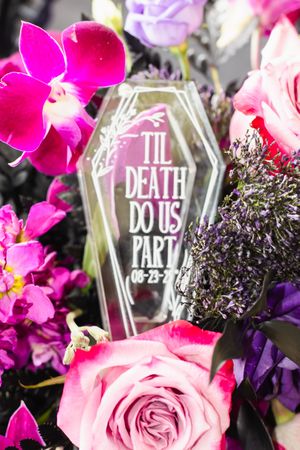 Gothic wedding decor featuring a coffin-shaped sign reading 'Til Death Do Us Part' surrounded by pink flowers.
