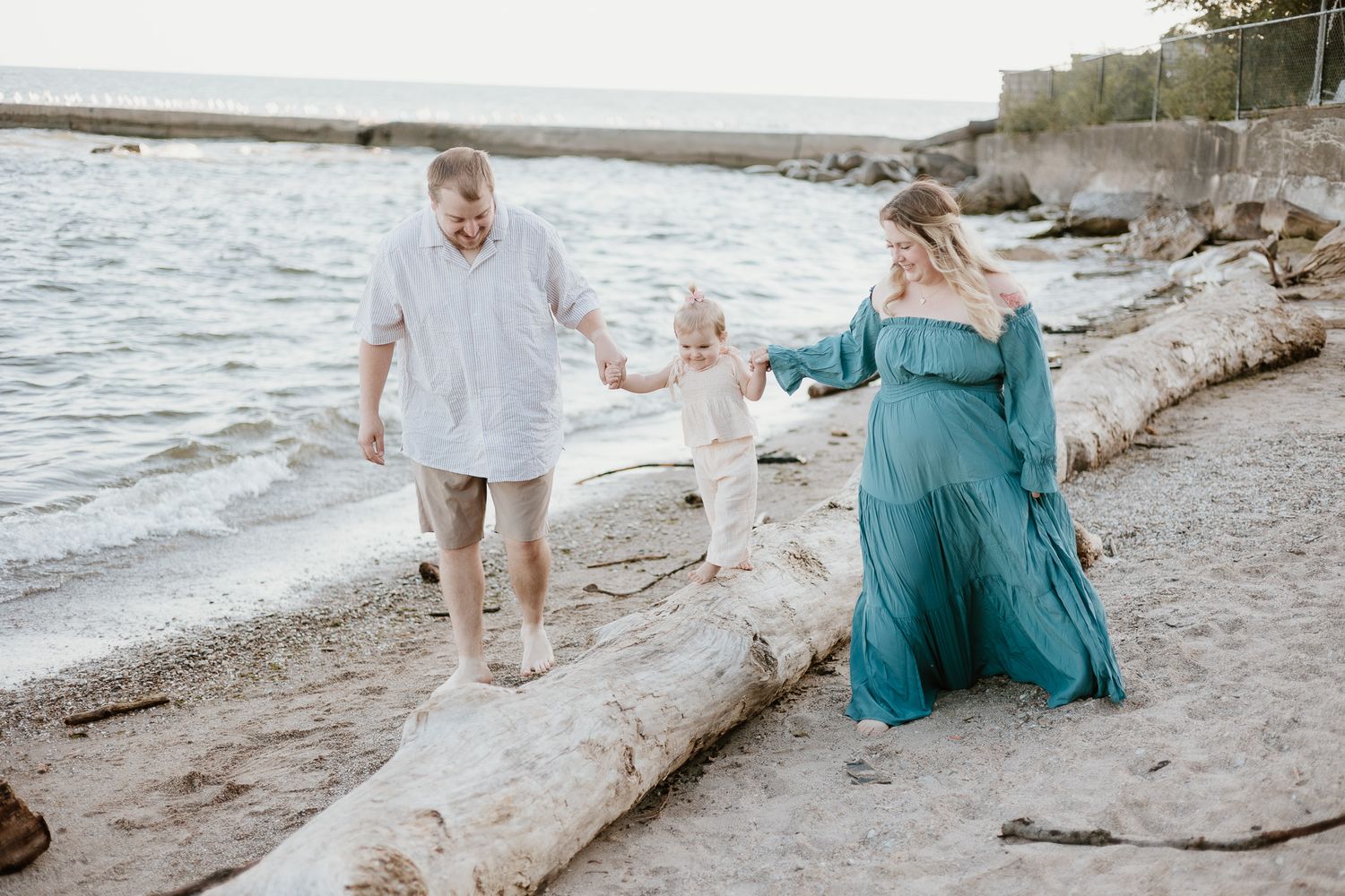 A man, woman, and young child are holding hands at the beach. The young child is balancing on a fallen tree in the sand.