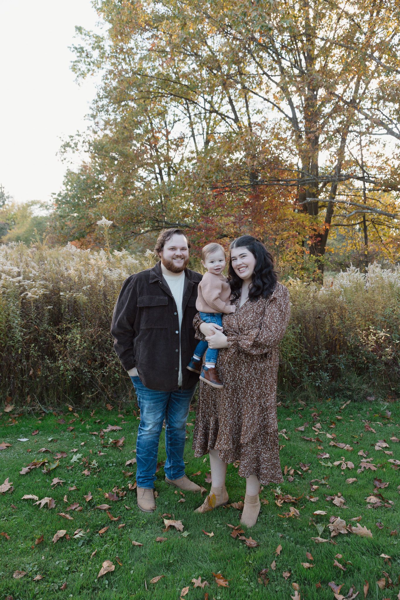 A family of three is standing outdoors in front of tall grass smiling