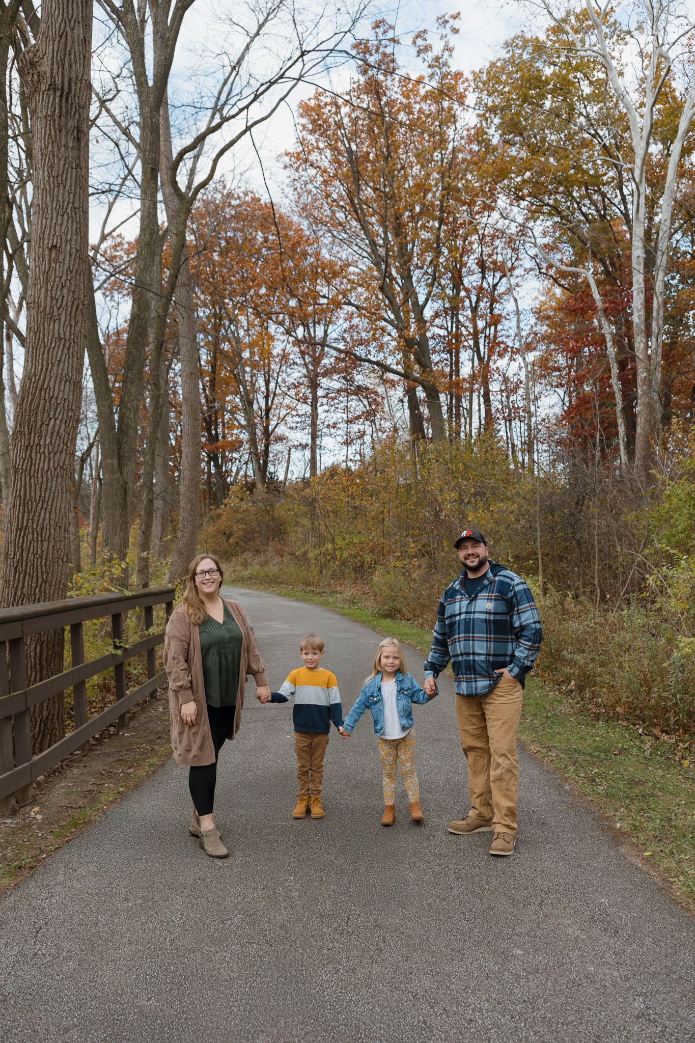 A family of four is holding hands, standing on a park bike path and smiling into the camera.