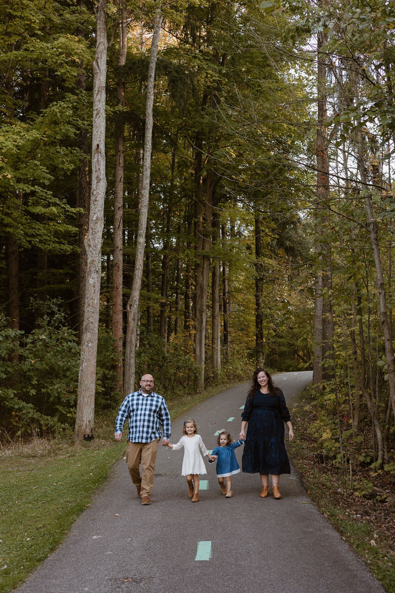 A family of four is holding hands, walking on a park bike path and smiling into the camera.