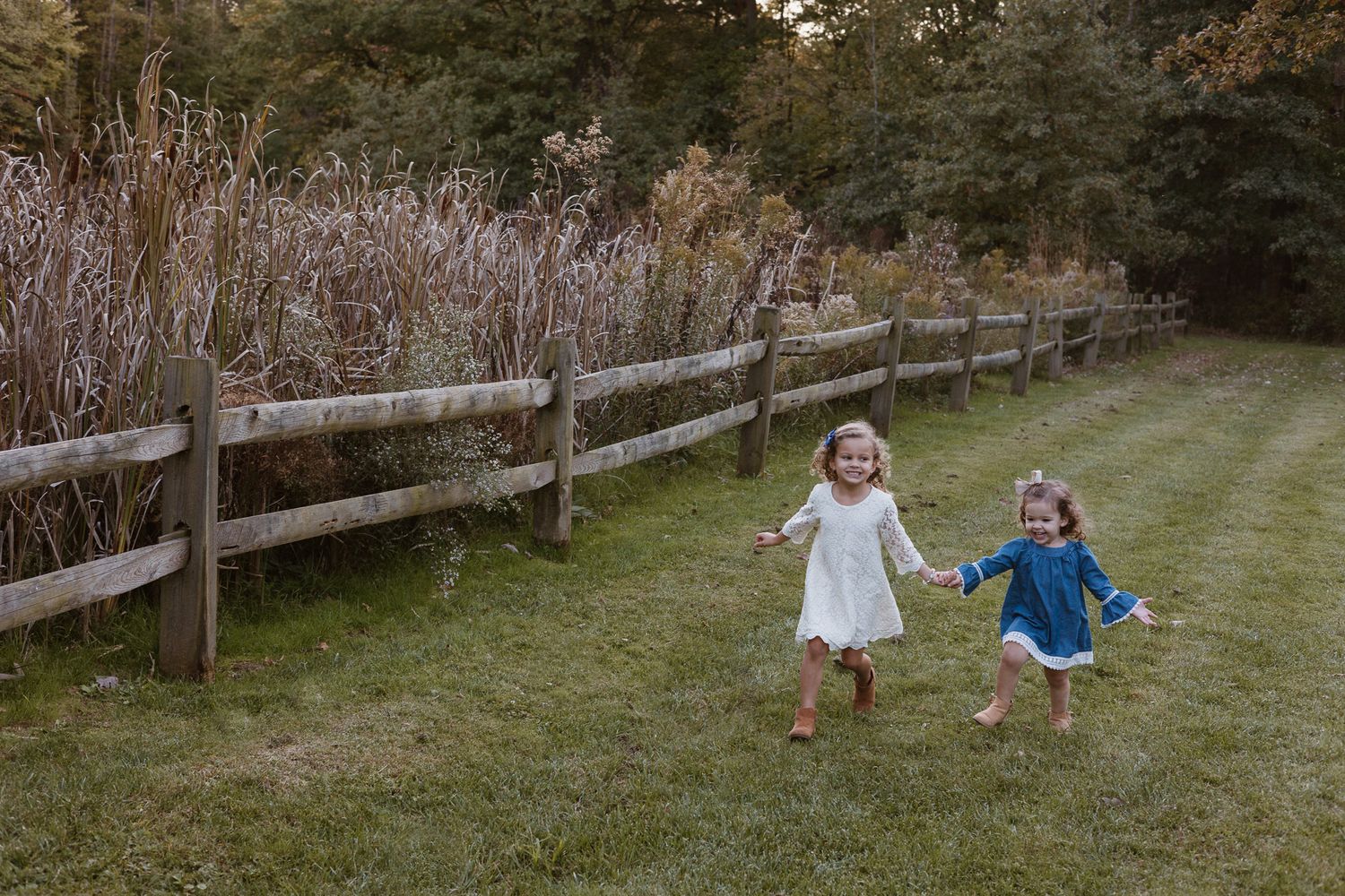 Two young girls hold hands and are running through grass towards the camera.