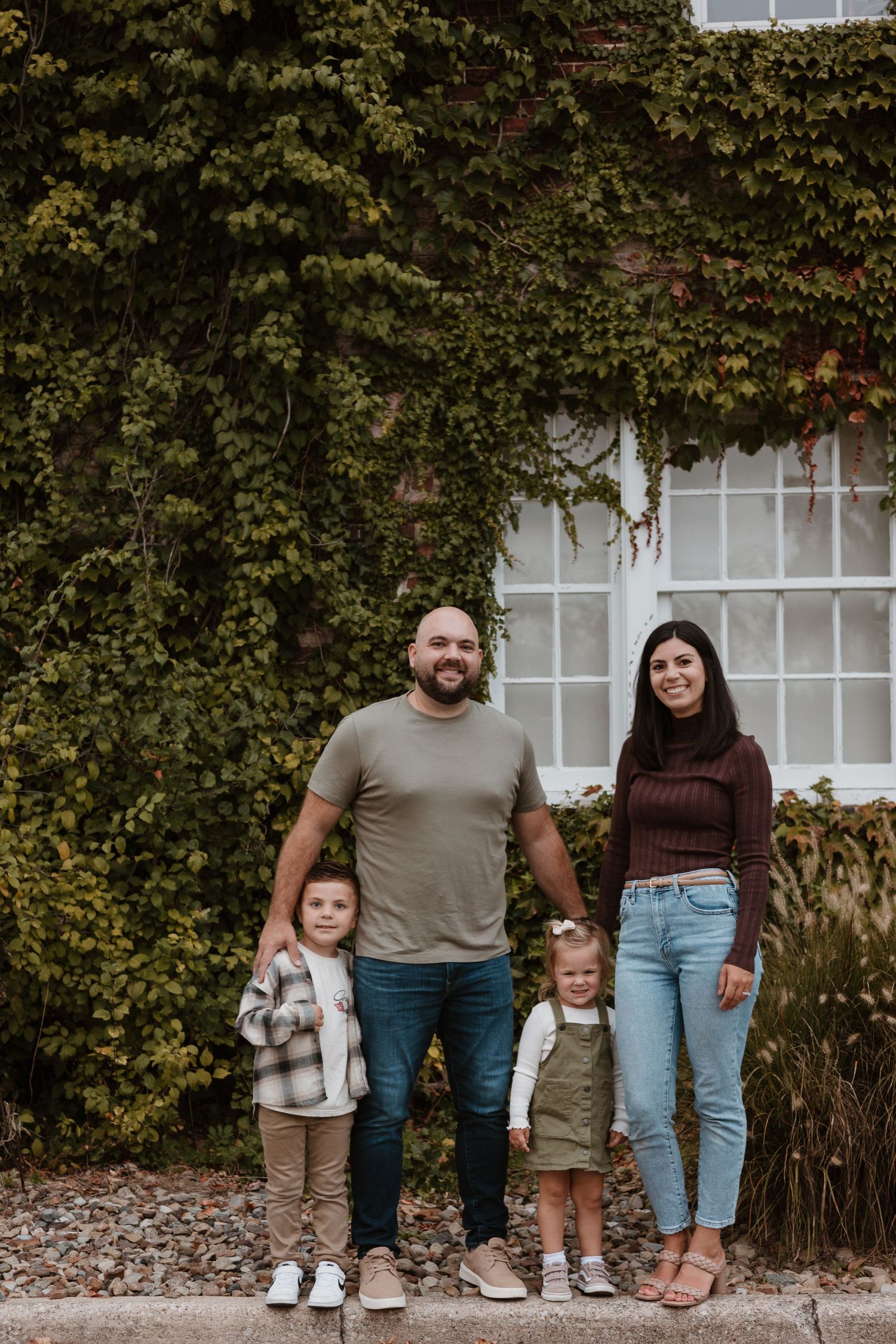 A family is standing in front of a building which has ivy growing on the exterior. A dad has his arms around his two young children.