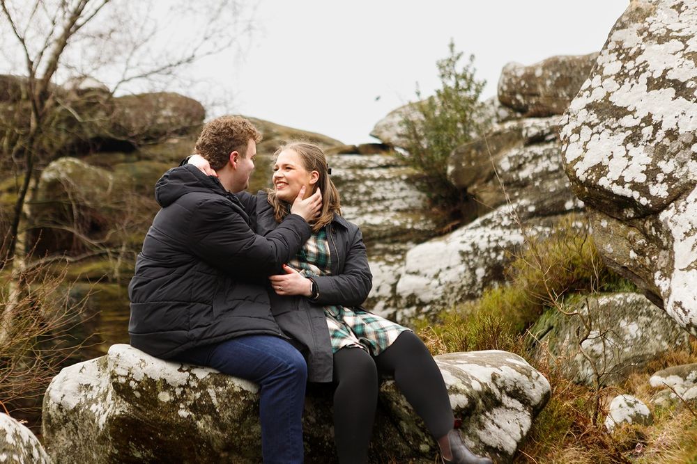 A couple embraces while sitting on a rocky outcrop in a scenic mountainous landscape.