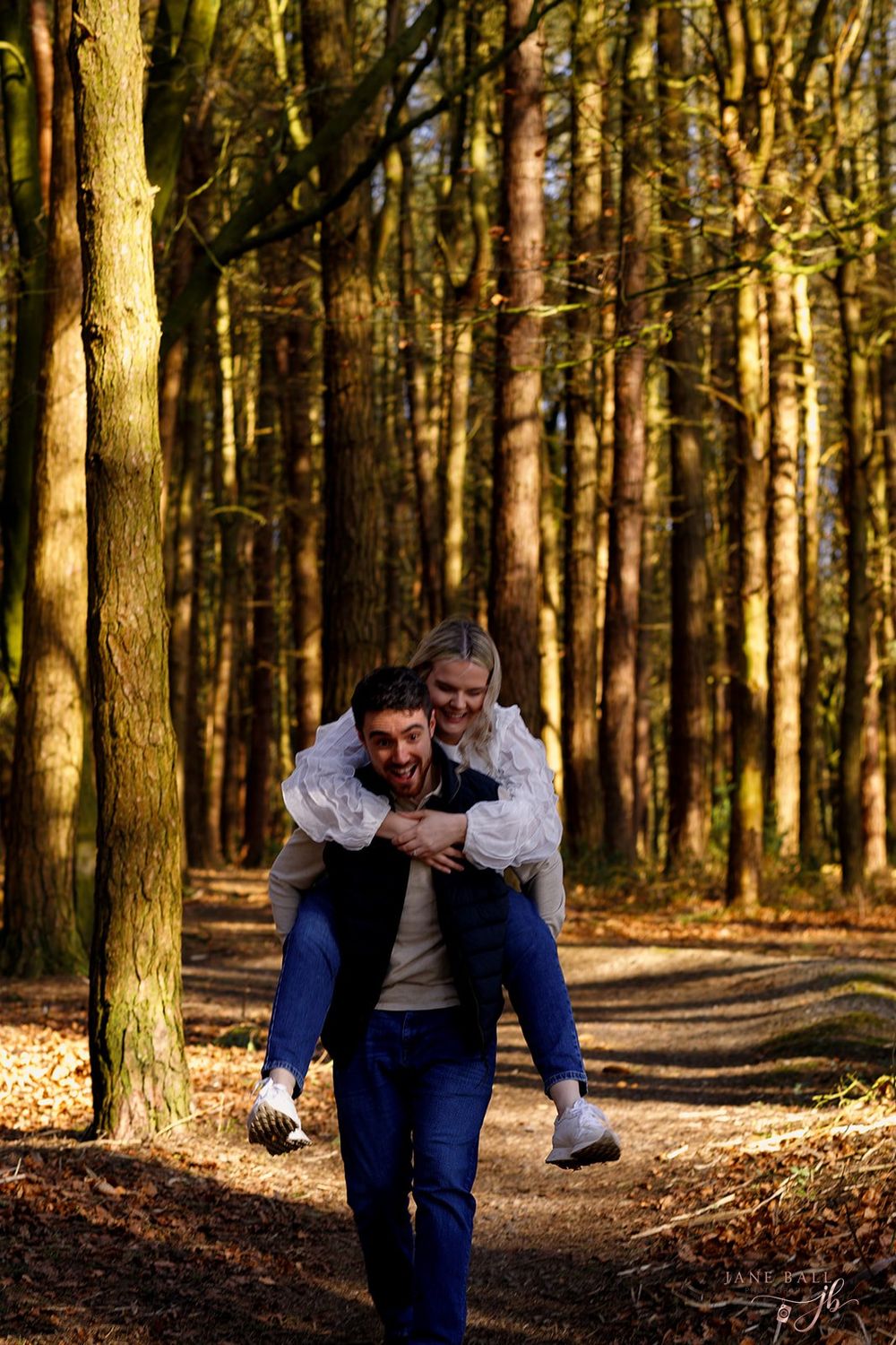 Two people playfully interact on a forest trail surrounded by tall trees in golden afternoon sunlight.