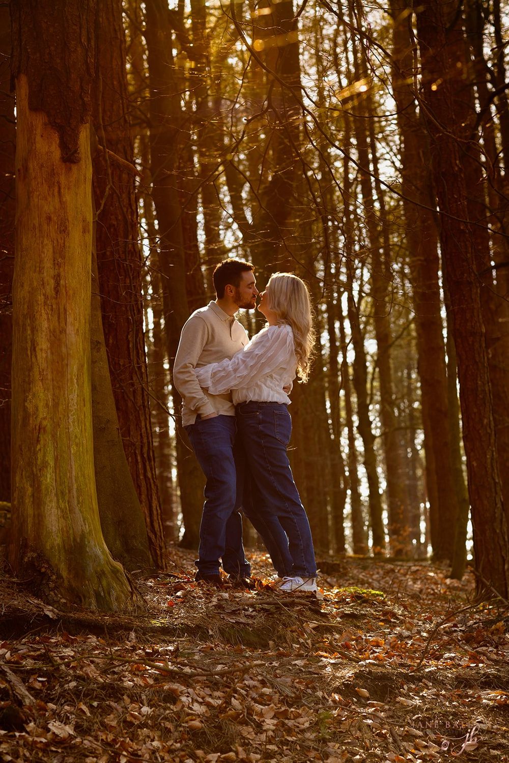 A couple shares a romantic moment among tall trees in a forest with fallen autumn leaves on the ground.