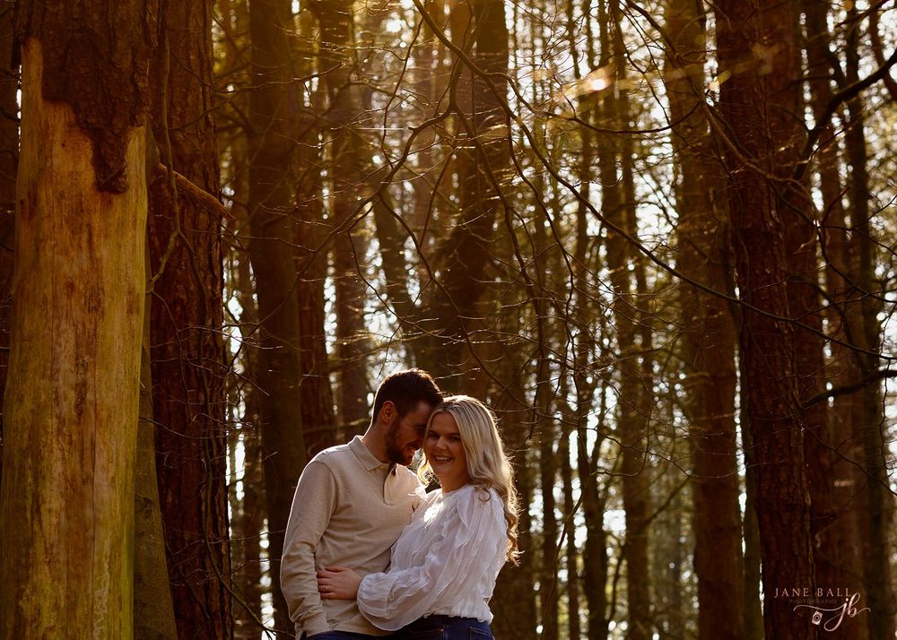 A romantic couple embraces in a sunlit forest with tall trees creating a warm, golden atmosphere.