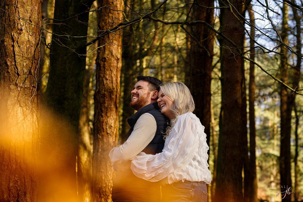 A romantic couple embraces in a sunlit forest with golden light filtering through tall pine trees.