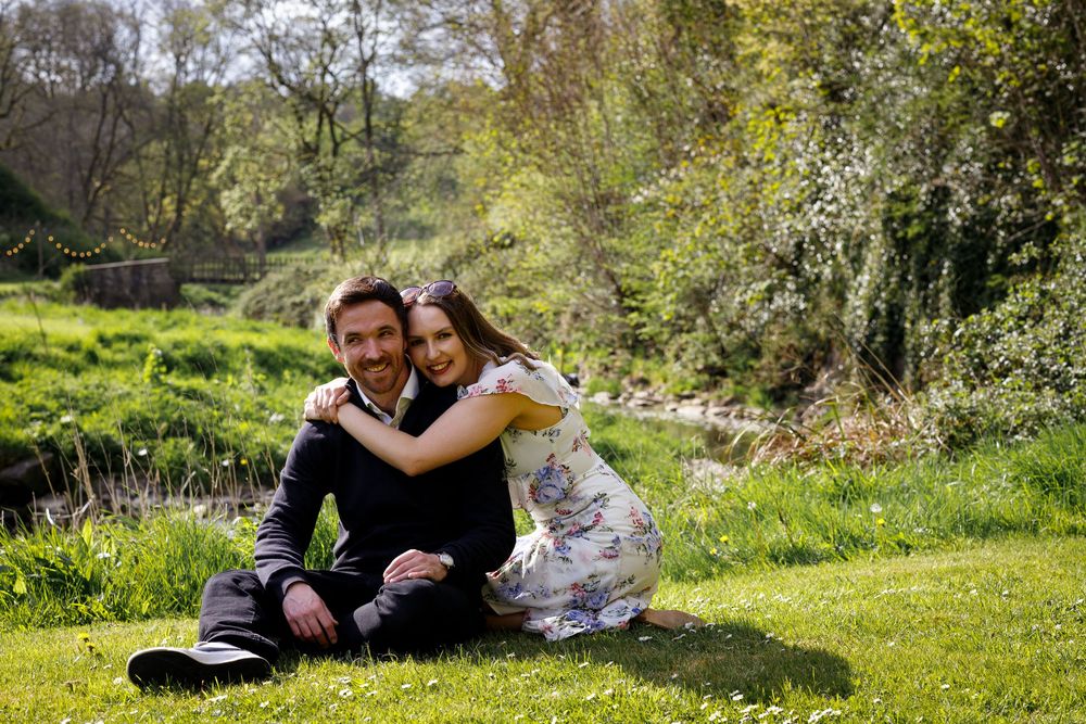 People sit and embrace in a sunlit garden surrounded by lush green vegetation and flowering bushes.