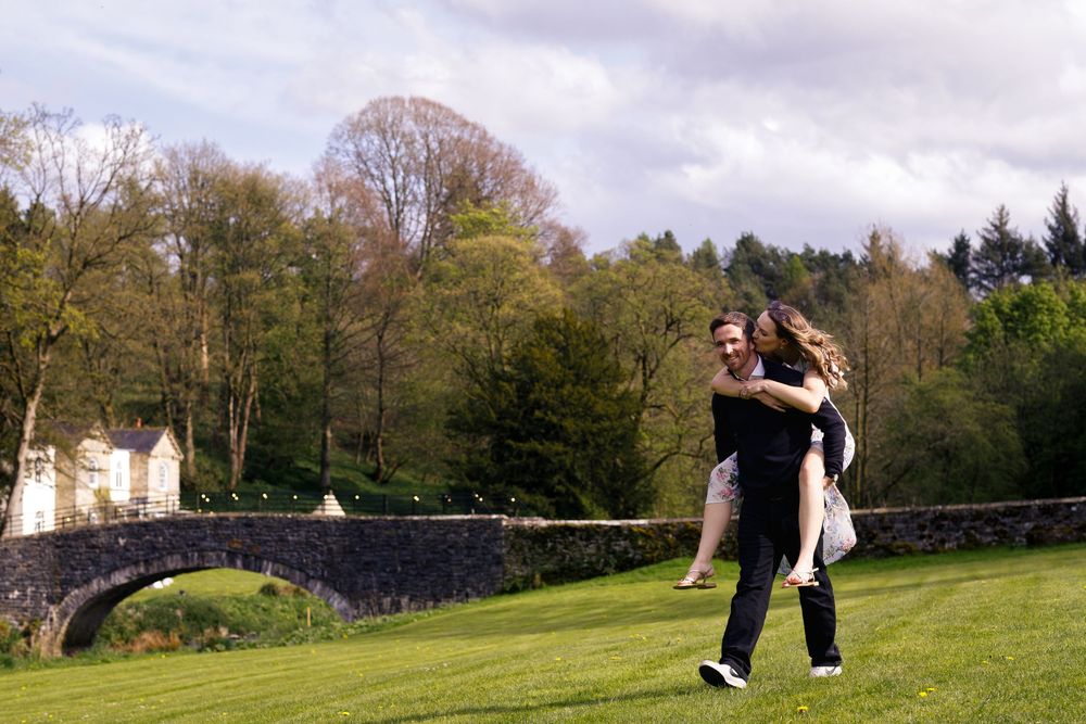 A couple in formal attire embrace while dancing on a green lawn near a stone bridge with trees in the background.