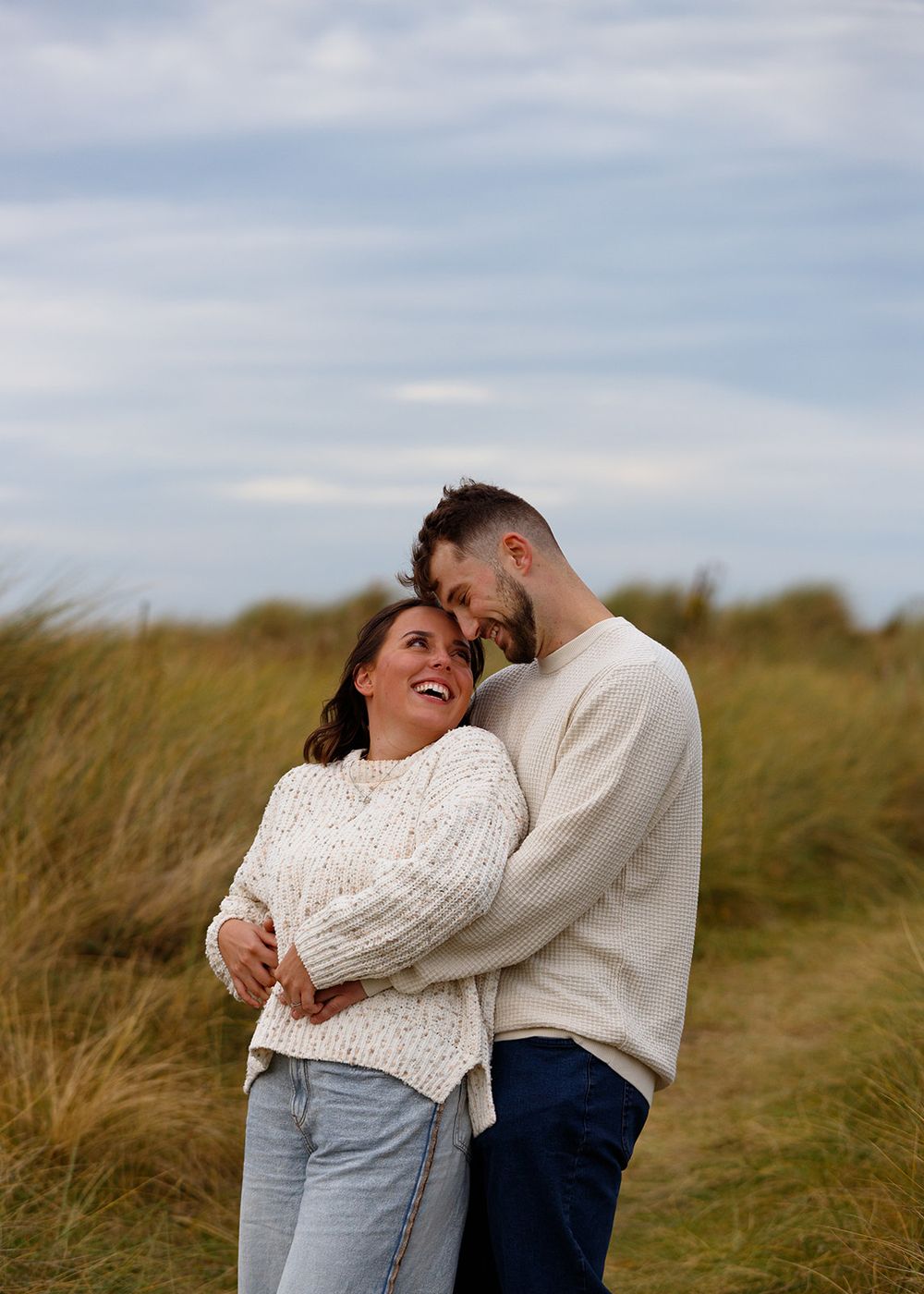 Candid moment between couple laughing in chunky sweaters among coastal grass.