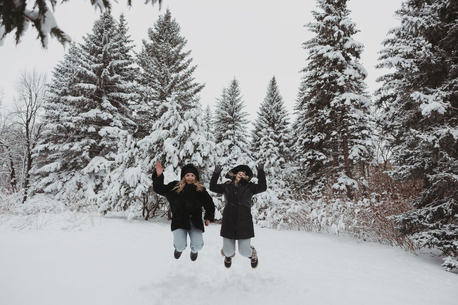 Two people jumping joyfully on a snowy path surrounded by snow-covered evergreen trees in winter.