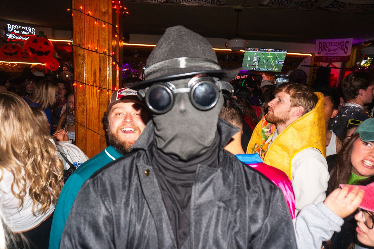 Someone in a dark gas mask costume poses at a crowded nightclub party with colorful lighting.
