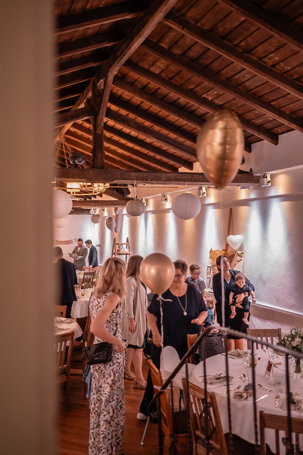 Guests mingle at an elegant indoor reception with exposed wooden beams, white tables and metallic balloon decorations.