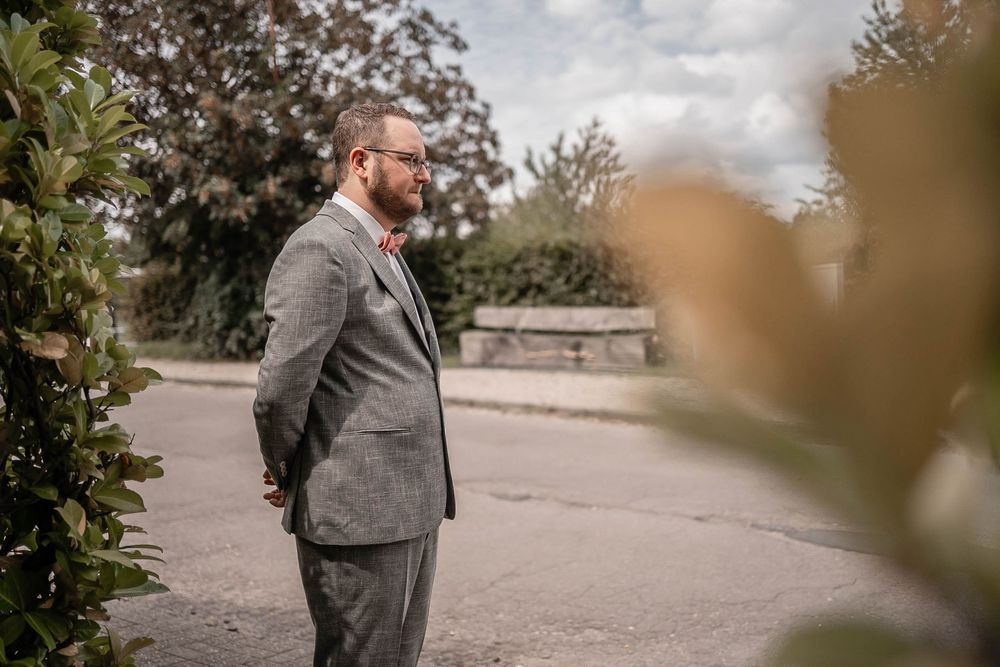 A person in a gray suit stands outdoors in an elegant garden setting during autumn.