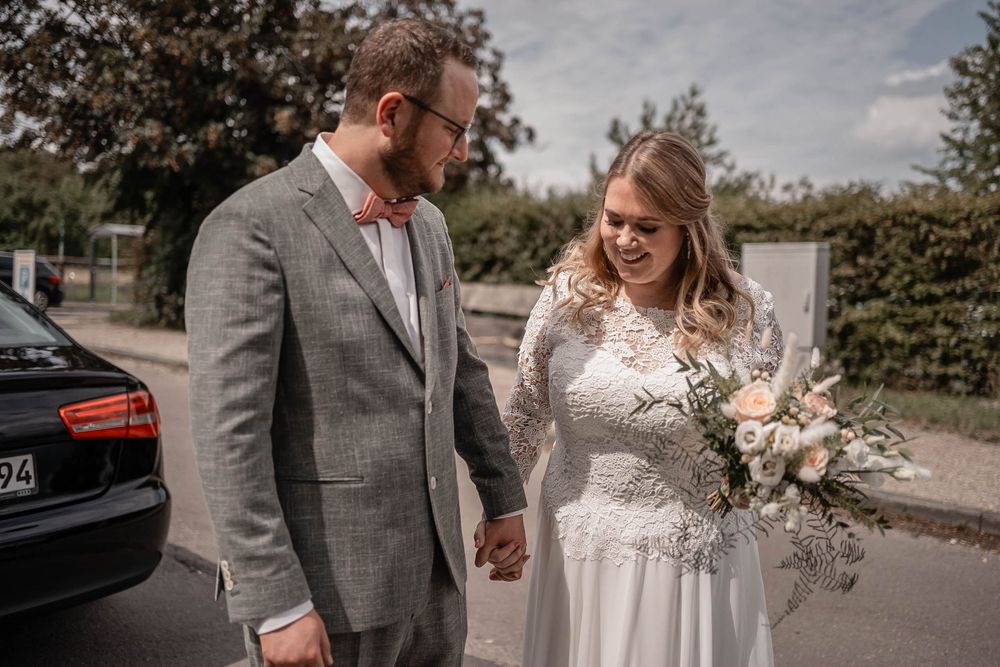 A couple in wedding attire standing next to a car, the bride wearing lace and holding a neutral-toned bouquet.