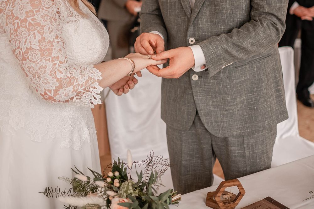 A couple's hands exchange wedding rings during a romantic outdoor ceremony with delicate greenery in the background.
