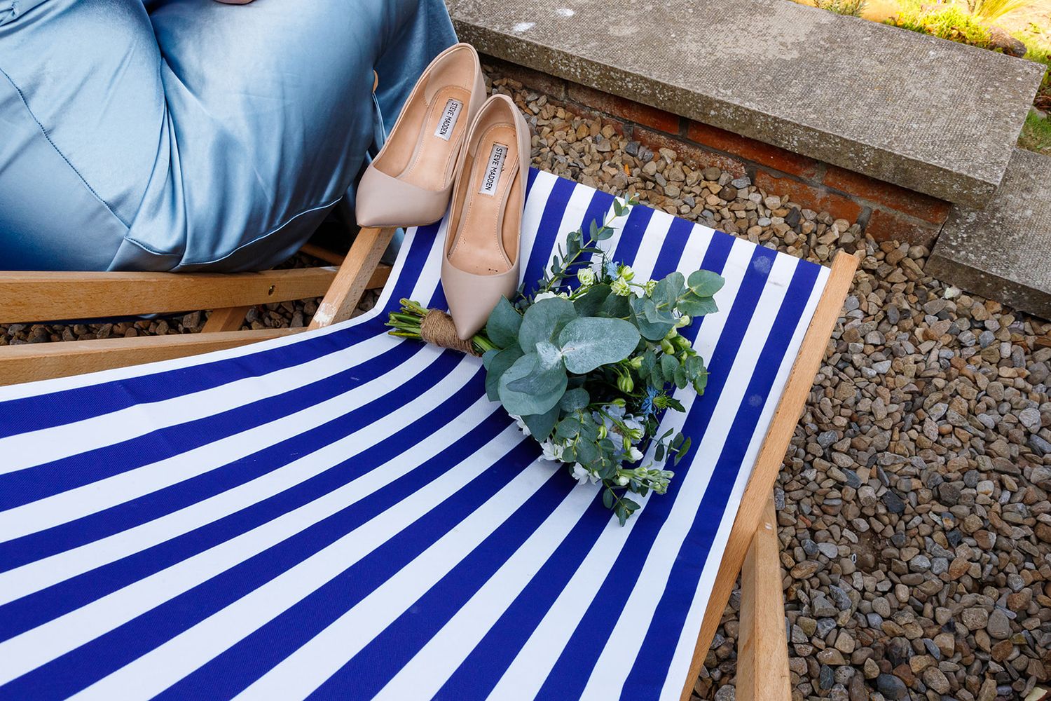 Nude heels and small floral bouquet resting on blue and white striped deck chair outdoors.
